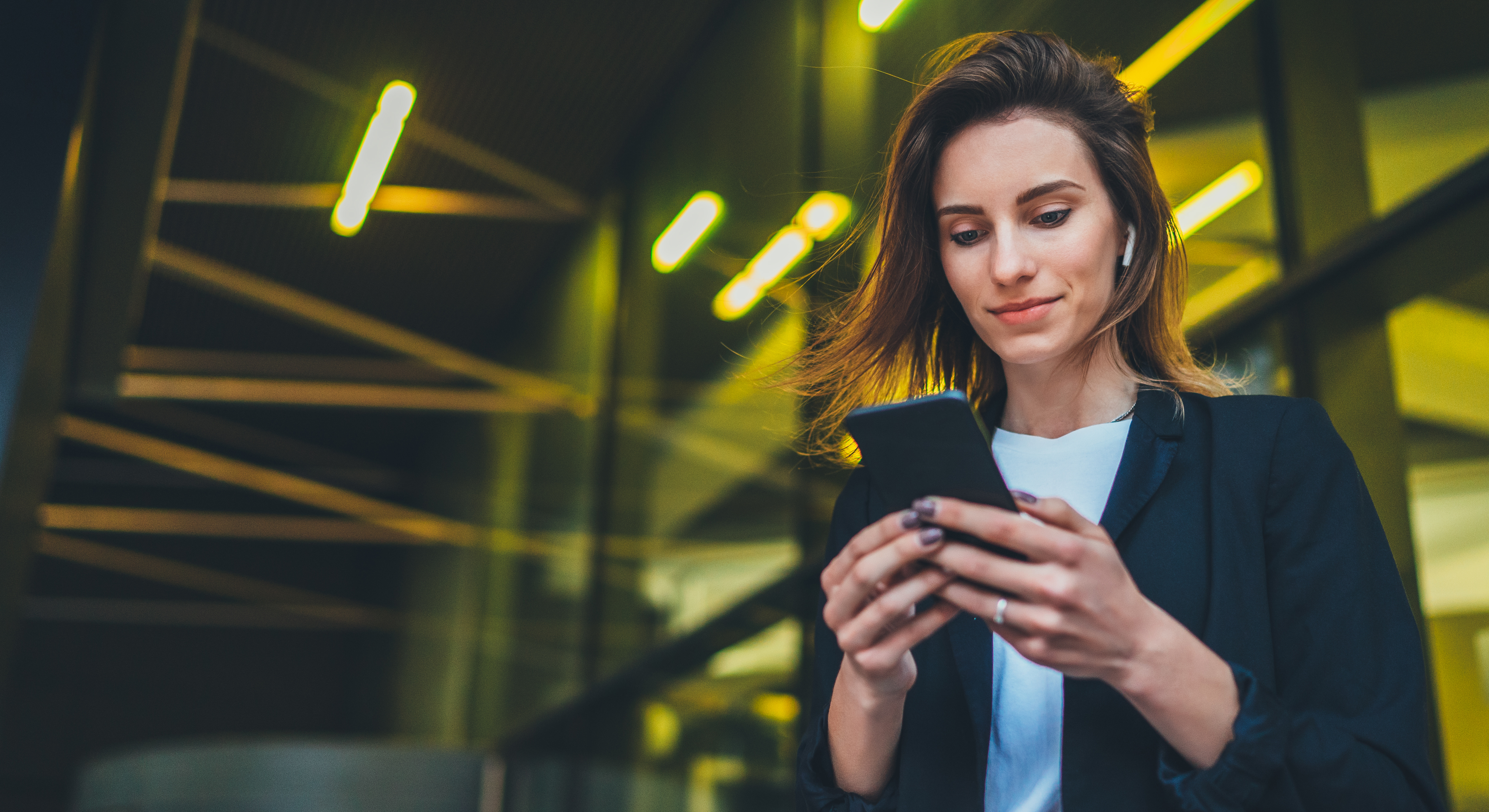 Woman wearing suit and Airpods looking at mobile phone with modern geometric lighting in background.