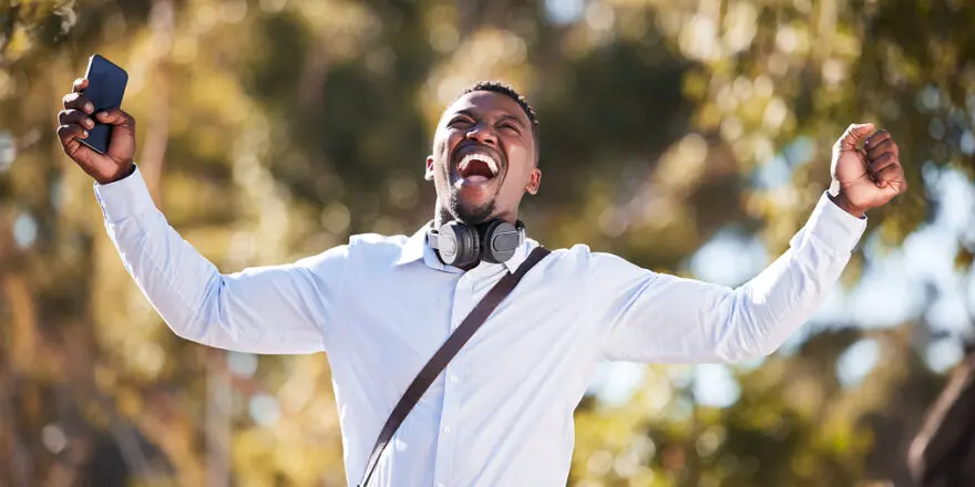 smiling black man in business attire outdoors with fancy headphones around his neck and leather cross body bag celebrating with arms up the freedom from monthly audiobook subscriptions