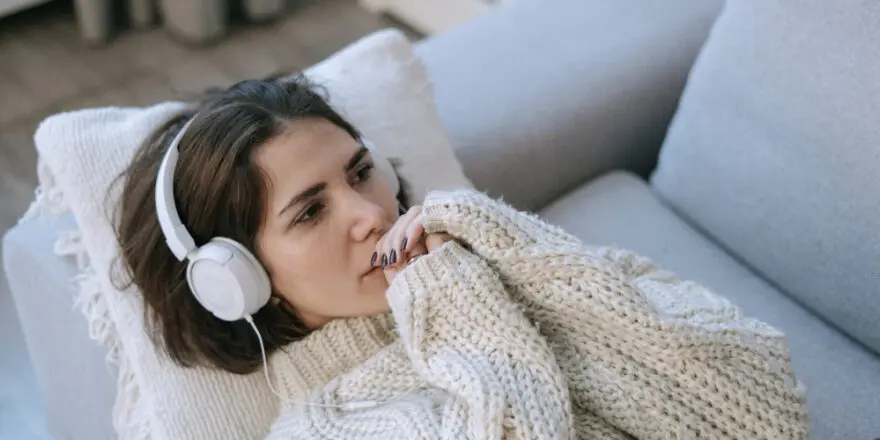 A woman wearing white headphones lies on a grey sofa wrapped in a chunky knit jumper, gazing thoughtfully into the distance while listening to something on her phone.