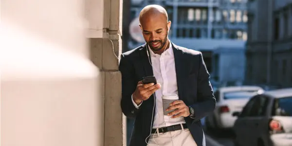 Man in a suit listening to xigxag audiobooks on his phone with wired earphones while standing on a city street, coffee cup in hand.