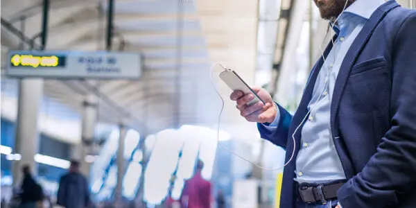 Man in a blazer standing on a train platform, listening to something on his phone with wired earphones.