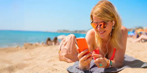 Woman lying on a sandy beach wearing orange sunglasses and earphones, smiling as she listens to an amazing audiobook on the xigxag app on her phone.