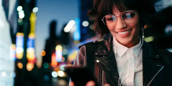 Young woman wearing glasses and a leather jacket listening to audiobooks and looking at the text on her phone with wired earphones standing in a modern cityscape at night.