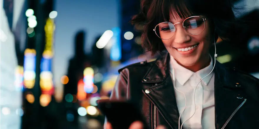Young woman wearing glasses and a leather jacket listening to audiobooks and looking at the text on her phone with wired earphones standing in a modern cityscape at night.