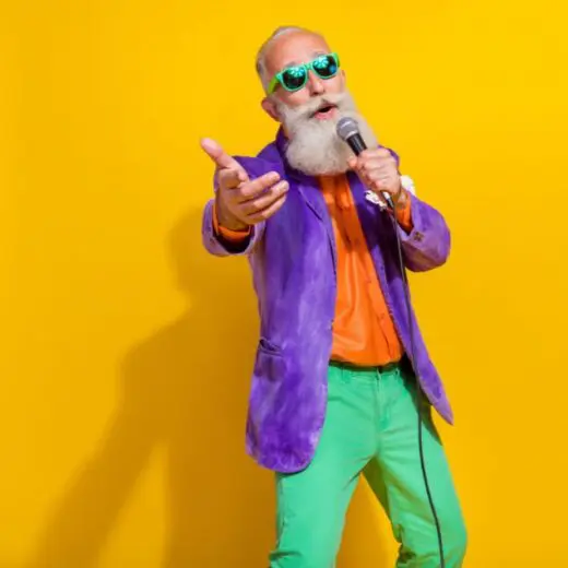Older man with a white beard wearing a purple jacket, orange shirt, green trousers and green sunglasses, speaking into a microphone against a bright yellow background.