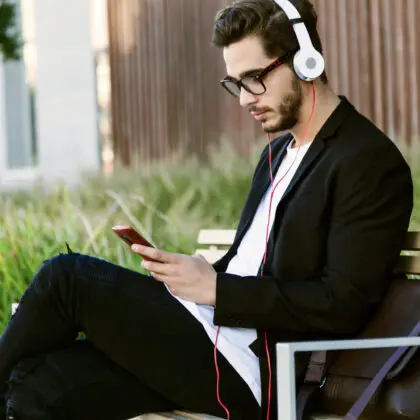 young man outside on a park bench, wearing headphones and looking at a smart phone in front of a lush green urban background