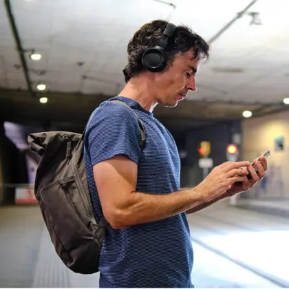 Man with rucksack on wearing headphones listening to audiobooks checking his phone at a train station