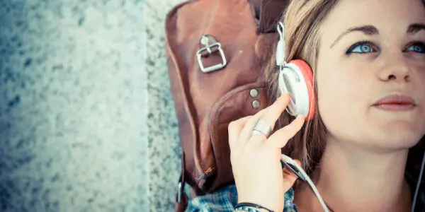 A young woman wearing headphones lies on the floor with her head resting against a leather travel bag, gazing upward as she listens to an audiobook.