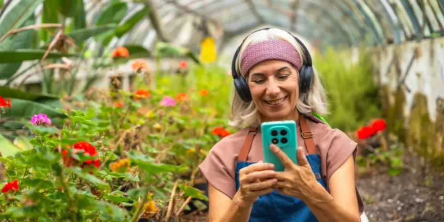 A woman wearing headphones and a pink patterned headband smiles as she enjoys an audiobook on xigxag and looks at her turquoise smartphone in a greenhouse. She is wearing a dusty pink t-shirt under a blue denim apron, surrounded by colourful flowers including geraniums and lantana.