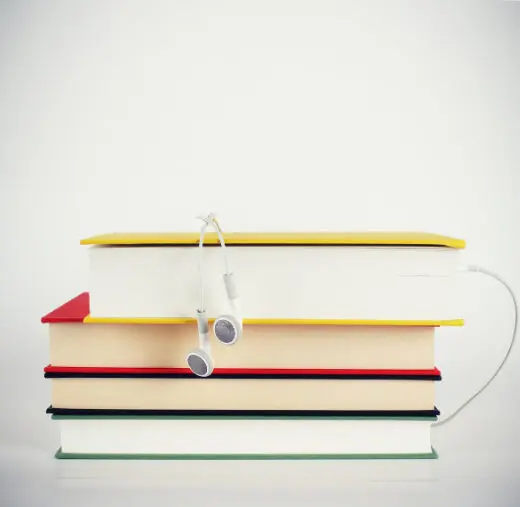 A stack of four hardback books with coloured spines (yellow, red and black) against a plain light grey background, with a pair of white earphones draped over the top book and a red bookmark poking out from the second.