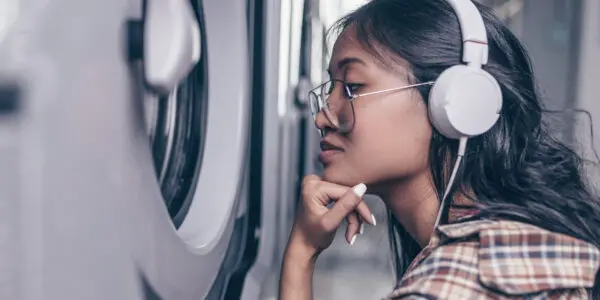 A young woman wearing glasses and white headphones listens to an audiobook while waiting for her laundry, chin resting on her hand.