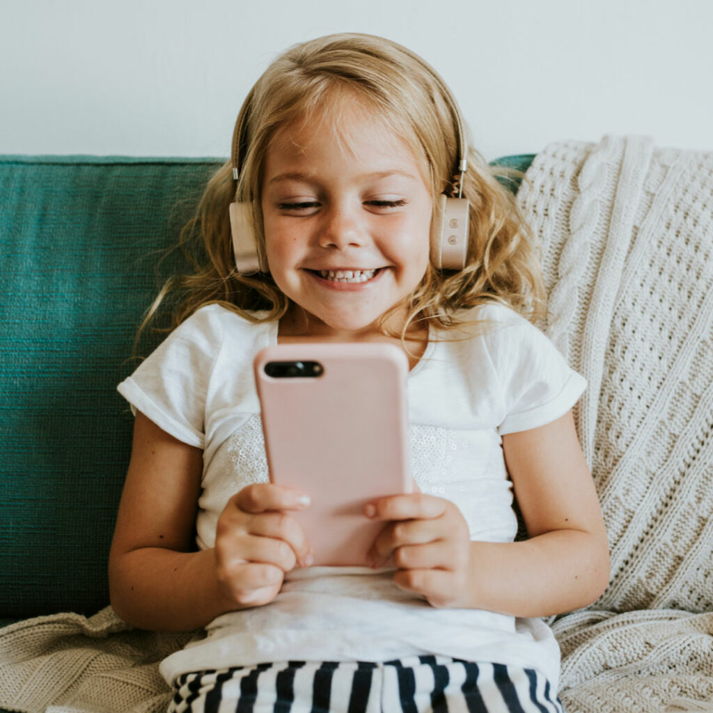 A young girl smiles while listening and reading a book on xigxag on a pink smartphone and matching headphones