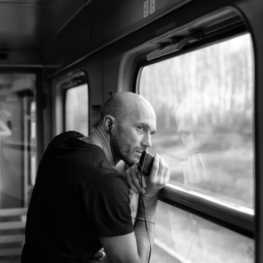 good looking man with a shaved head and headphones, holding a smartphone on a train looking out the window, listening to an audiobook on xigxag